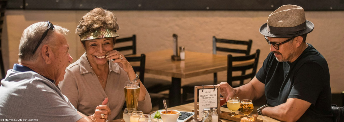 Drei lächelnde Menschen sitzen an einem Tisch mit Speisen und Getränken