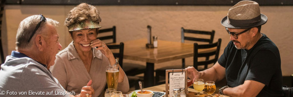 Drei lächelnde Menschen sitzen an einem Tisch mit Speisen und Getränken